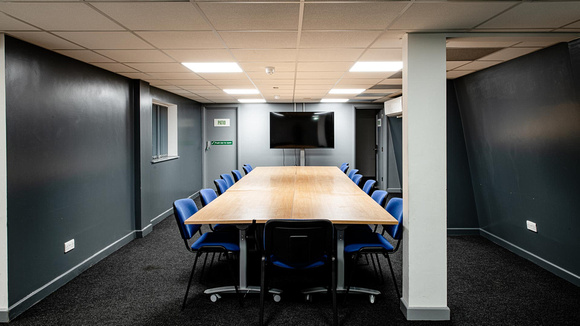 A room with a large table with 13 chairs around it. There is also a tv placed on the wall