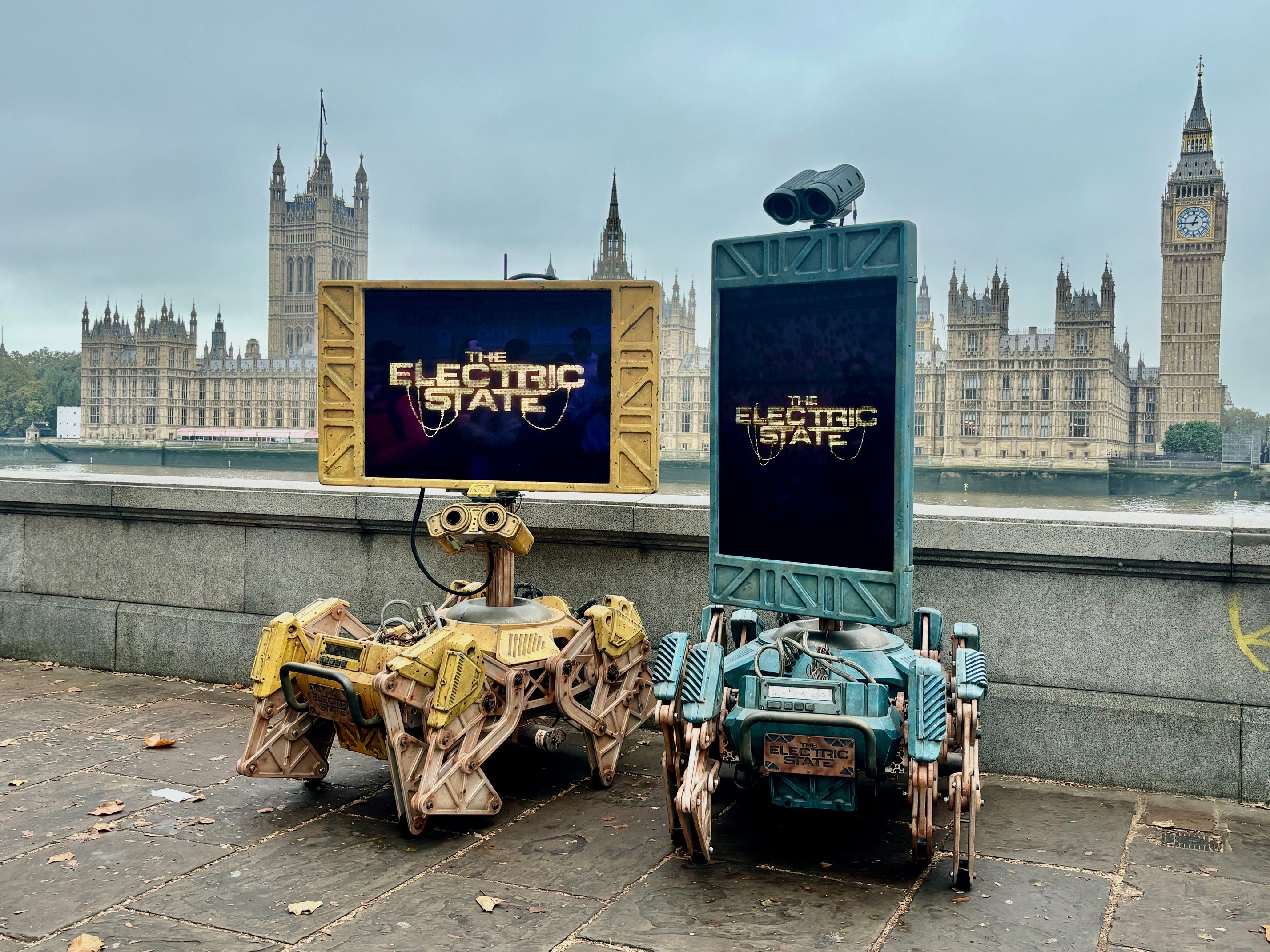 2 robots on Albert Embankment with the Houses of Parliament in the background. The robots show the message 'The Electric State'