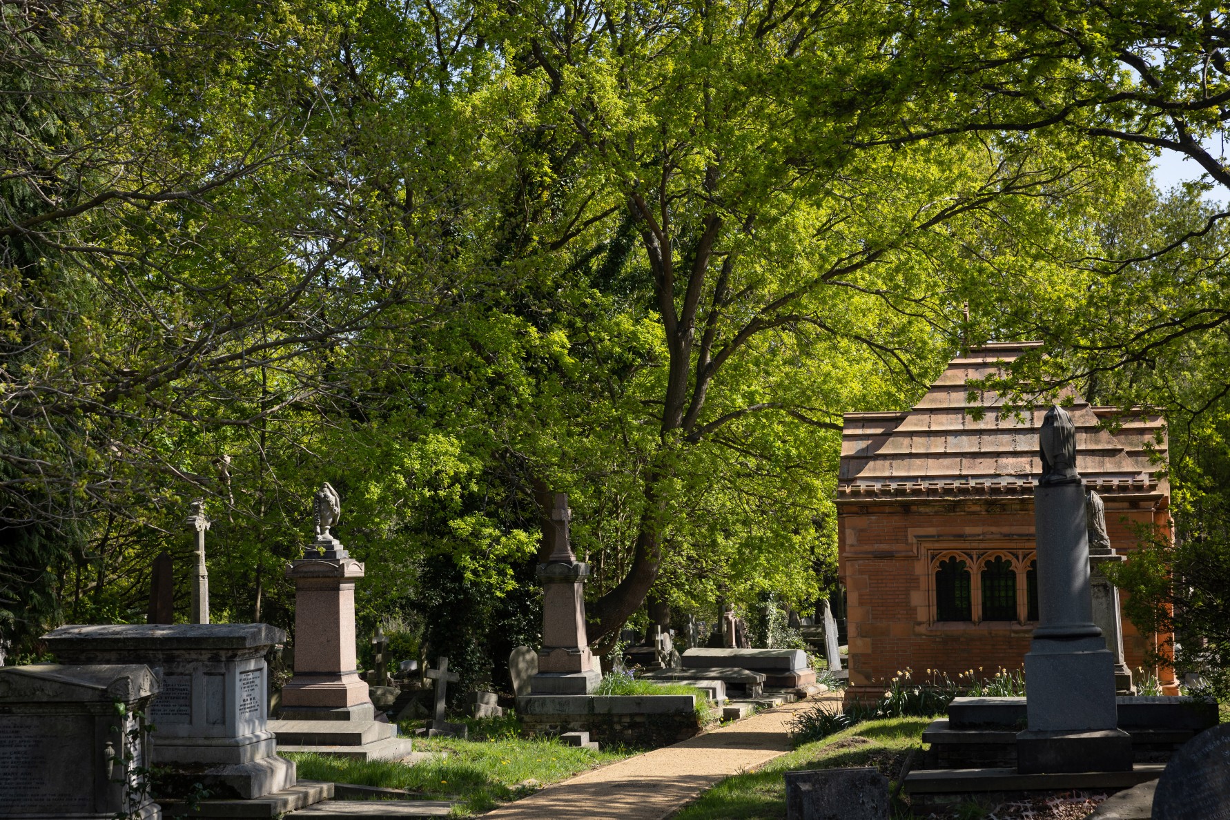 Tombstones and a small mausoleum in West Norwood Cemetery surrounded by lush green trees.